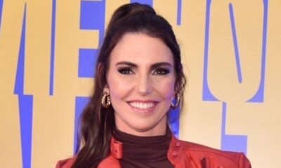 Glenda Kozlowski sorrindo, com brincos dourados e jaqueta vermelha, fundo amarelo e azul