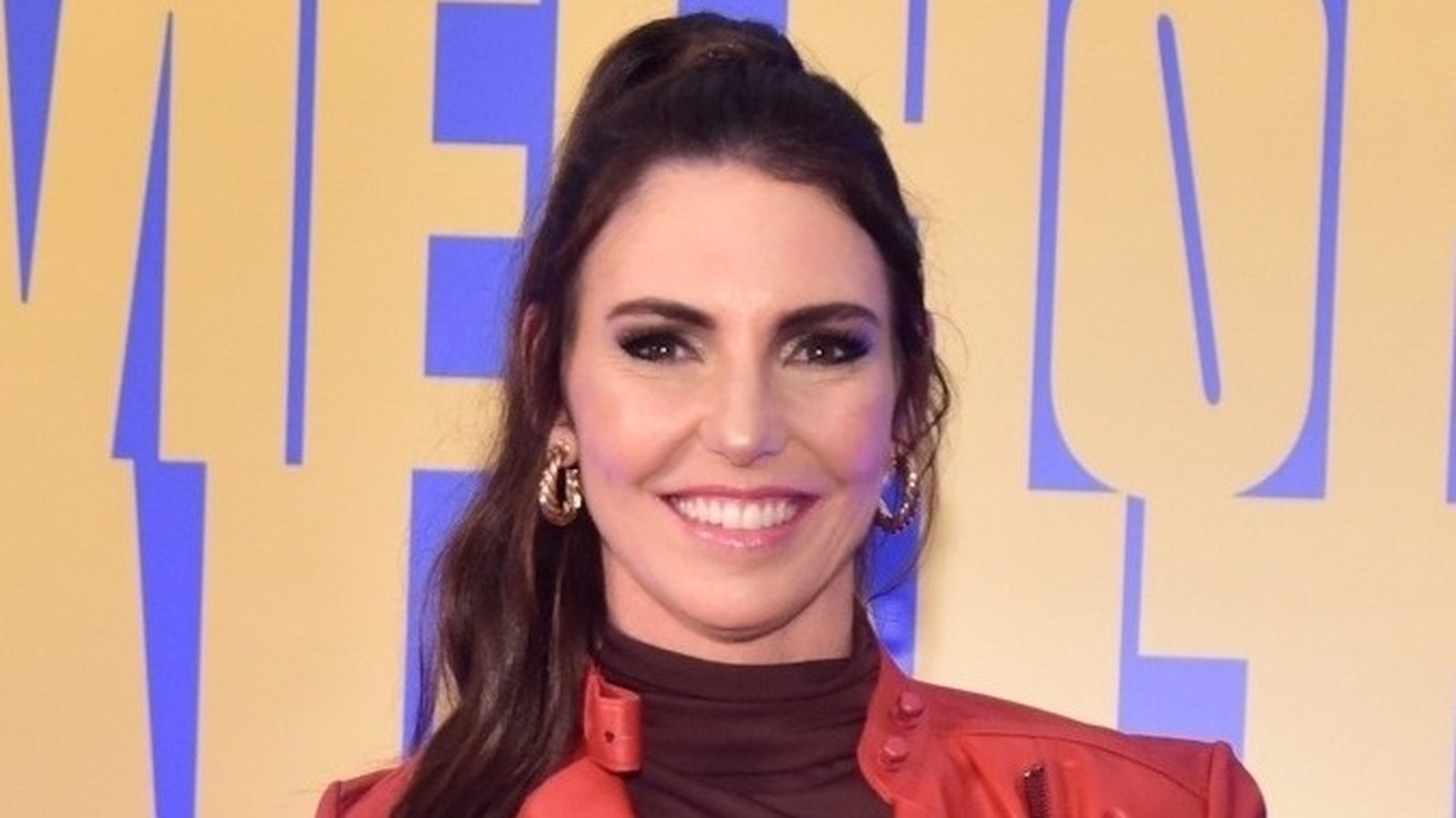 Glenda Kozlowski sorrindo, com brincos dourados e jaqueta vermelha, fundo amarelo e azul