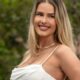 Yasmin Brunet sorrindo, cabelo loiro, colar dourado e blusa branca, fundo verde desfocado.