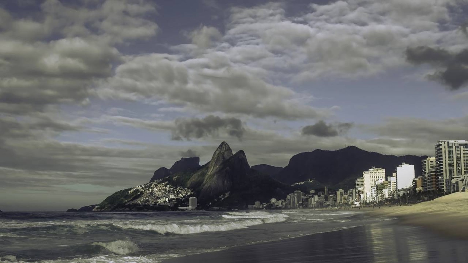 Praia com ondas, prédios da cidade e montanhas sob céu nublado no Rio de Janeiro