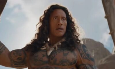 Dwayne Johnson como Maui, cabelos longos, tatuagens corporais, colar de dentes e expressão séria.