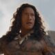 Dwayne Johnson como Maui, cabelos longos, tatuagens corporais, colar de dentes e expressão séria.