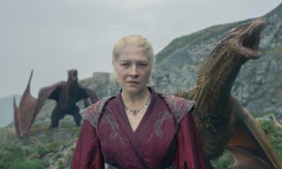 Mulher loira em vestes vermelhas olha à frente, com dois dragões gigantes em paisagem rochosa.