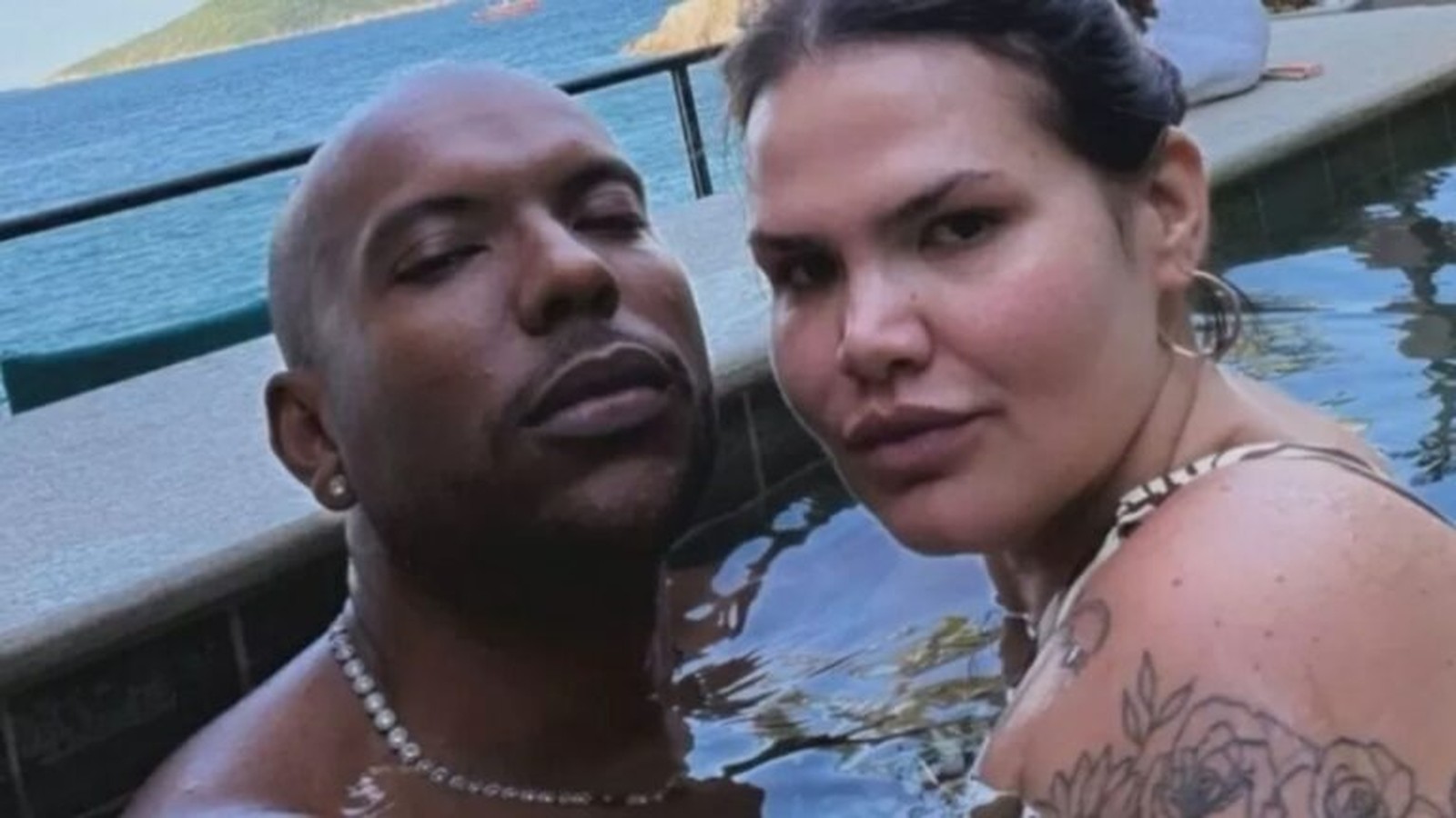 Homem careca e mulher com tatuagens submersos em piscina, olhando para câmera
