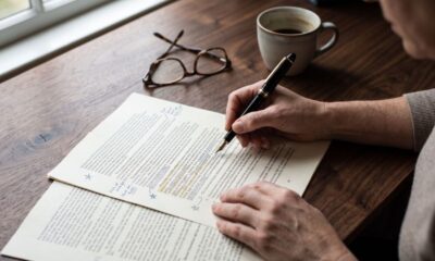 Pessoa revisa documentos com caneta tinteiro, óculos e xícara de café em mesa de madeira
