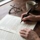 Pessoa revisa documentos com caneta tinteiro, óculos e xícara de café em mesa de madeira