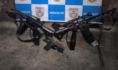 Dois fuzis, um com "QUITUNGO" no corpo, uma pistola e rádio em chão de concreto, fundo PMERJ