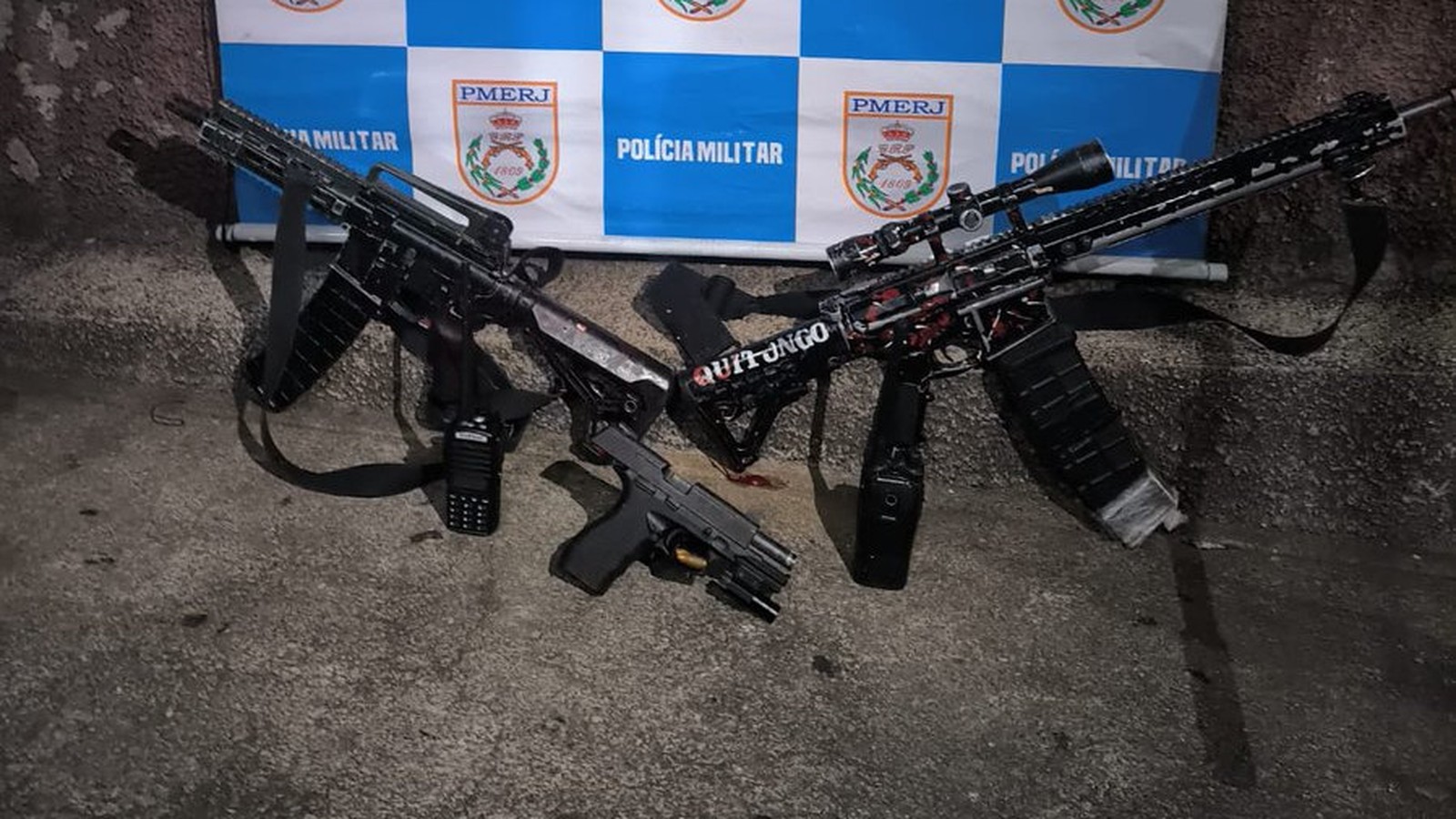 Dois fuzis, um com "QUITUNGO" no corpo, uma pistola e rádio em chão de concreto, fundo PMERJ