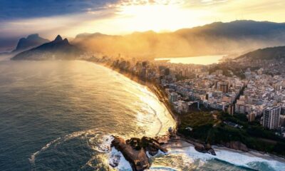 Vista aérea de cidade costeira com praia, prédios, montanhas e névoa dourada ao sol