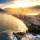 Vista aérea de cidade costeira com praia, prédios, montanhas e névoa dourada ao sol