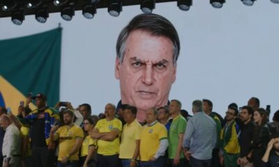 Tela exibe rosto bravo de Bolsonaro a público com celulares, bandeira do Brasil visível