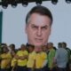 Tela exibe rosto bravo de Bolsonaro a público com celulares, bandeira do Brasil visível