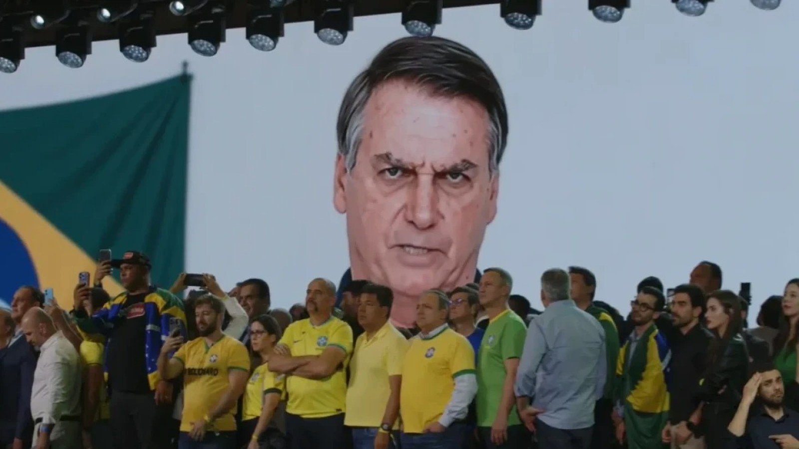 Tela exibe rosto bravo de Bolsonaro a público com celulares, bandeira do Brasil visível