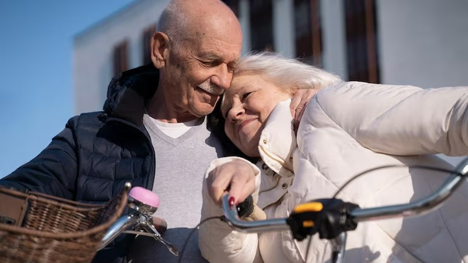 Casal de idosos abraçado com carinho ao lado de uma bicicleta com cesta, sob céu azul