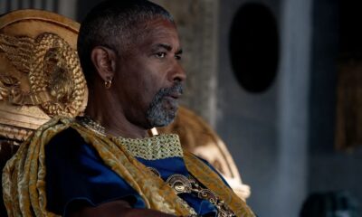 Denzel Washington em perfil com barba grisalha, vestindo túnica azul e dourada, sentado em trono