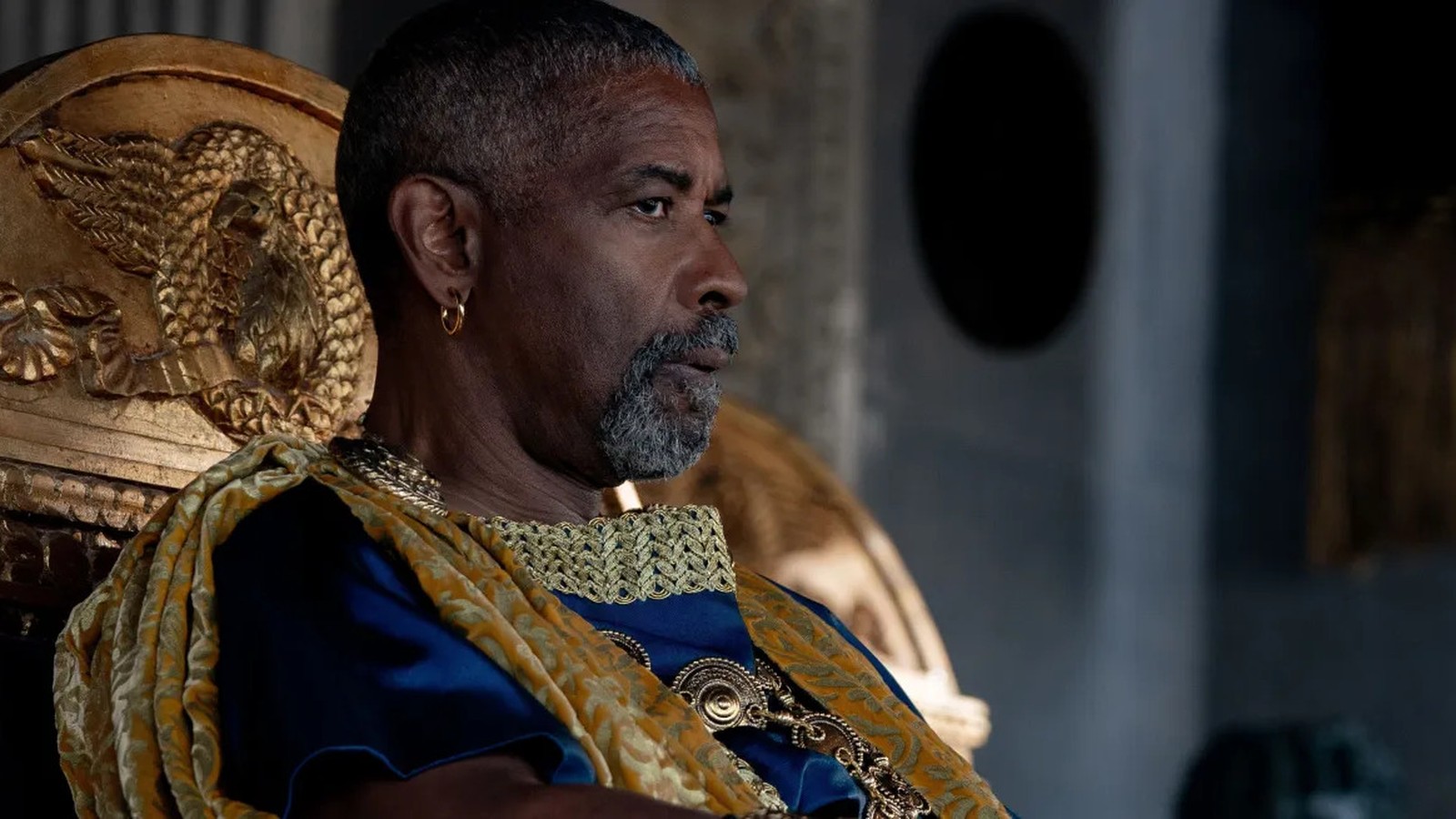 Denzel Washington em perfil com barba grisalha, vestindo túnica azul e dourada, sentado em trono