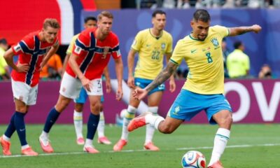 Jogador brasileiro de camisa amarela chuta a bola em campo; jogadores noruegueses de vermelho observam.