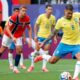 Jogador brasileiro de camisa amarela chuta a bola em campo; jogadores noruegueses de vermelho observam.