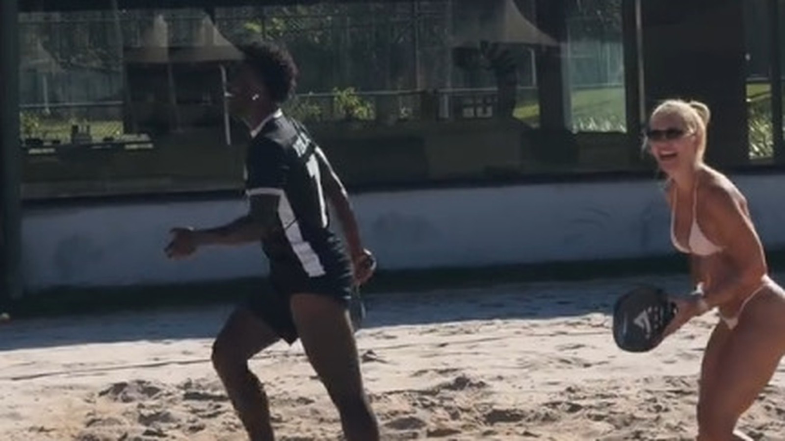 Virginia Fonseca de biquíni sorrindo, com raquete de beach tennis, Vini Jr. correndo na areia