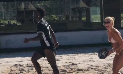 Virginia Fonseca de biquíni sorrindo, com raquete de beach tennis, Vini Jr. correndo na areia