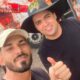 Selfie de dois homens sorrindo, um de boné e outro com chapéu preto escrito CAJU #N012