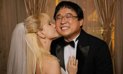 Mulher loira de véu beija homem de óculos e terno preto em foto de casamento