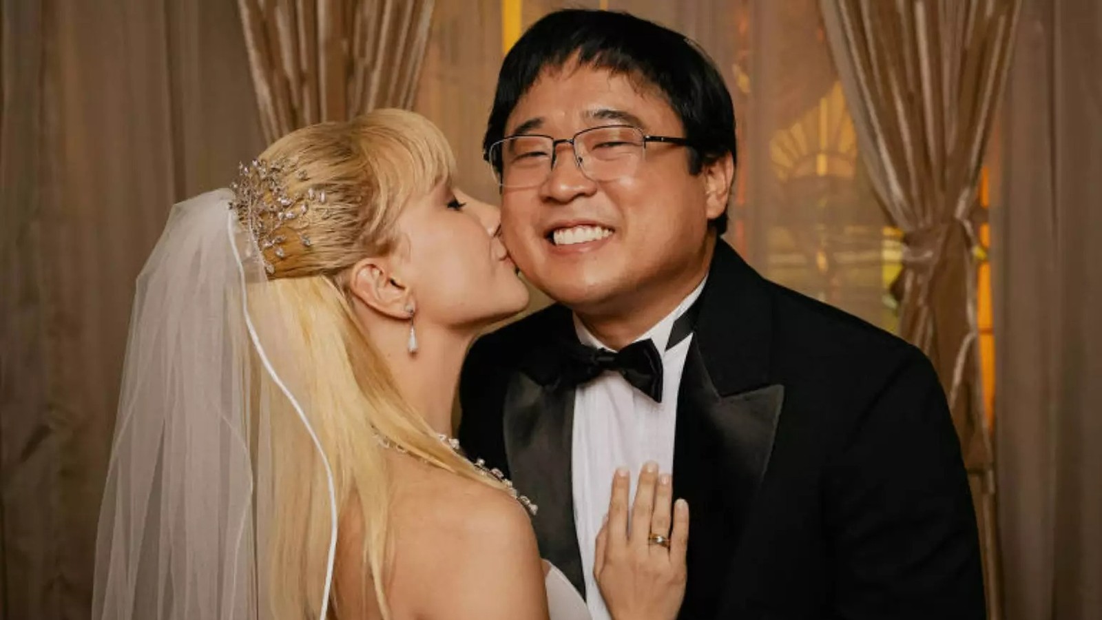 Mulher loira de véu beija homem de óculos e terno preto em foto de casamento