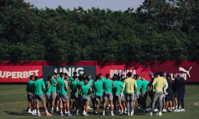 Jogadores de futebol em huddle no campo de treino, com placa SUPERBET e árvores