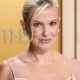 Close-up de Millie Bobby Brown sorrindo levemente, cabelo loiro preso, brincos, fundo desfocado com NETFLIX
