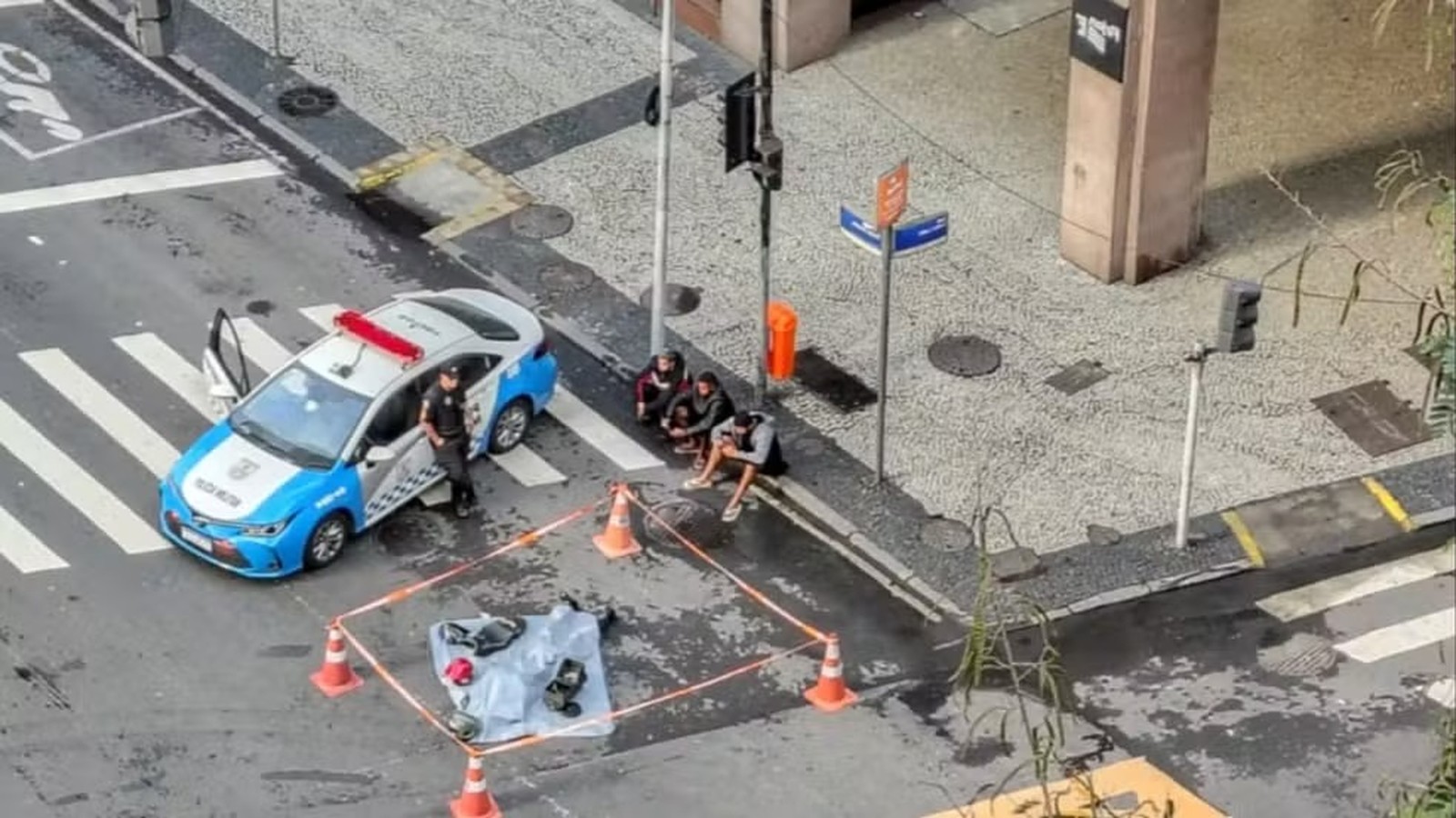 Viatura policial, policial, três homens sentados na calçada e cena de acidente isolada com cones na rua