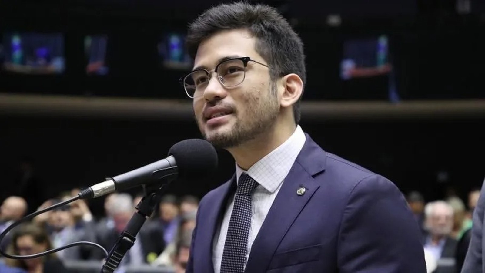 Kim Kataguiri de óculos, camisa xadrez e gravata, discursa ao microfone em plenário