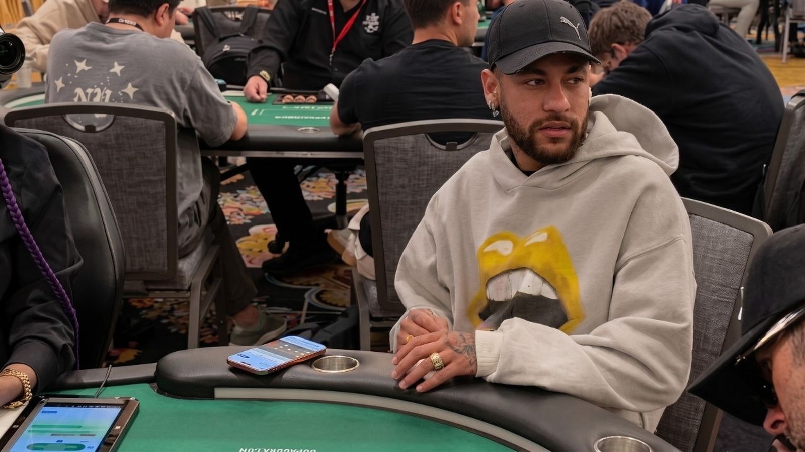 Neymar de boné e moletom cinza com estampa amarela, sentado em mesa de pôquer