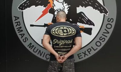 Homem de costas, algemado, camisa preta, logo de águia e textos "ARMAS MUNIÇ EXPLOSIVOS"
