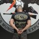 Homem de costas, algemado, camisa preta, logo de águia e textos "ARMAS MUNIÇ EXPLOSIVOS"