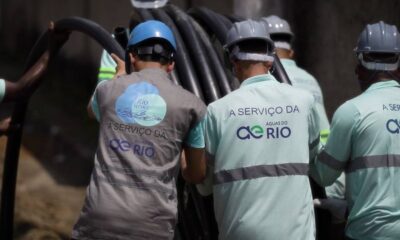 Trabalhadores com capacetes e uniformes 'Águas do Rio' e 'Rio Service' manuseiam tubos