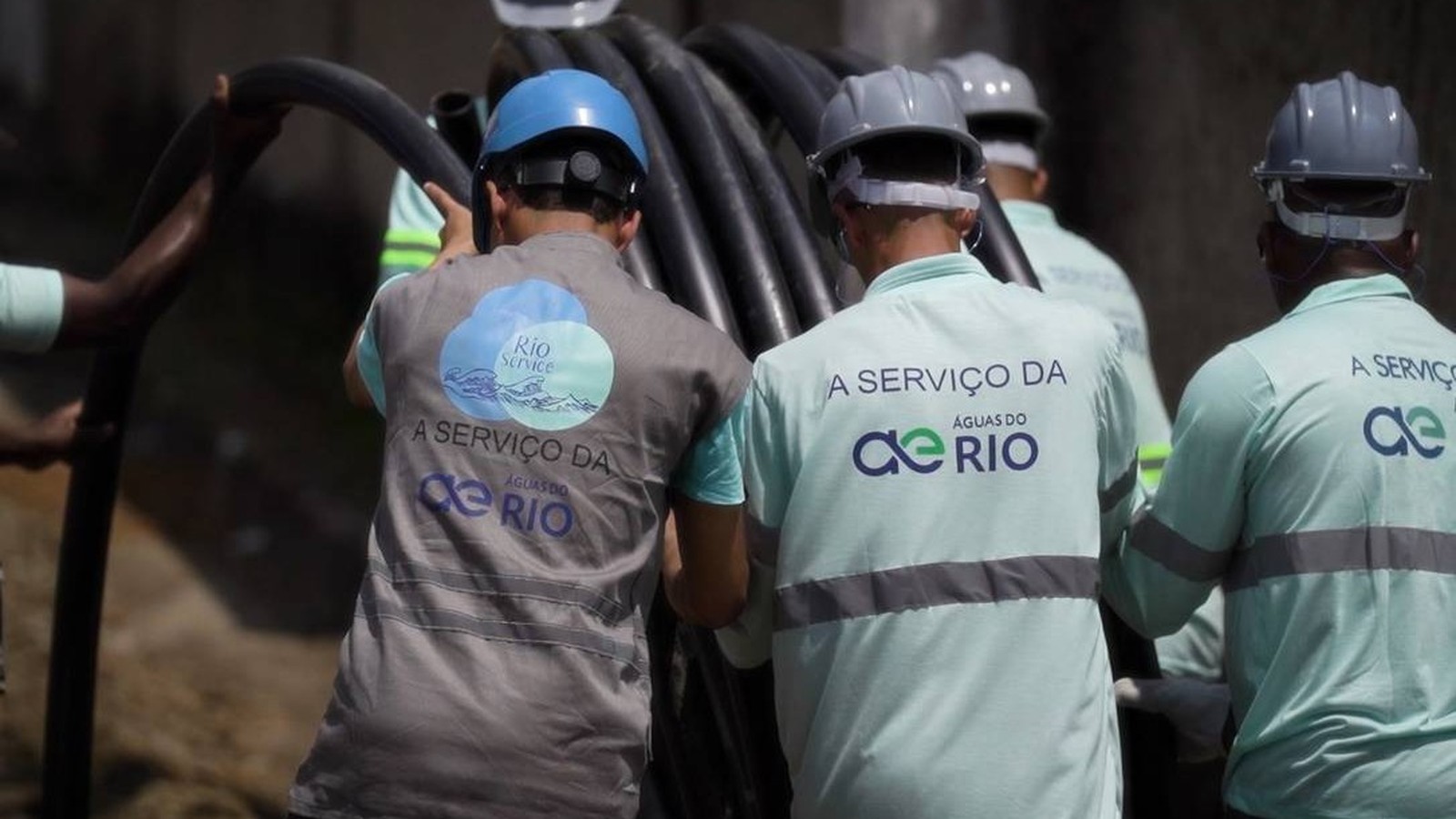 Trabalhadores com capacetes e uniformes 'Águas do Rio' e 'Rio Service' manuseiam tubos