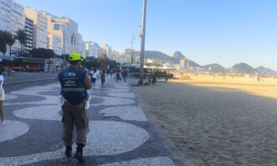 Agente da Ordem Pública com colete azul e boné amarelo caminha no calçadão da praia de Copacabana.