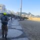 Agente da Ordem Pública com colete azul e boné amarelo caminha no calçadão da praia de Copacabana.