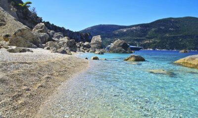 Praia de pedras com mar azul-turquesa cristalino, rochas, montanha e barco ao fundo