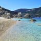 Praia de pedras com mar azul-turquesa cristalino, rochas, montanha e barco ao fundo