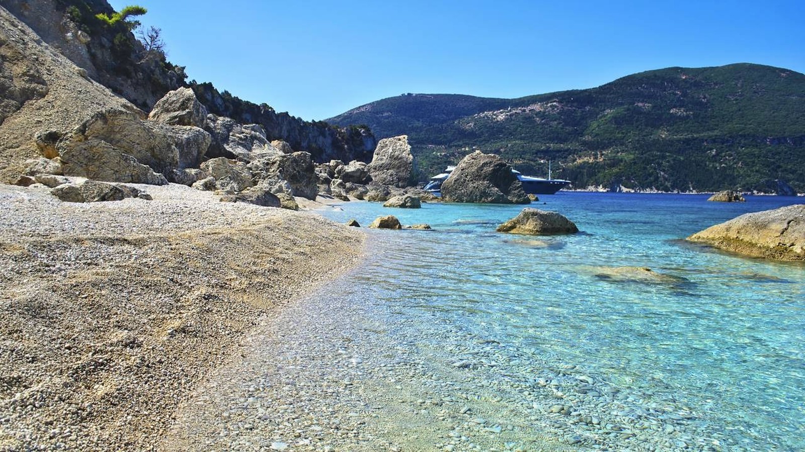 Praia de pedras com mar azul-turquesa cristalino, rochas, montanha e barco ao fundo