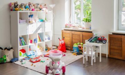 Quarto infantil desorganizado com brinquedos no chão, prateleira branca cheia de bichos de pelúcia e janela.