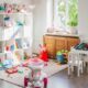 Quarto infantil desorganizado com brinquedos no chão, prateleira branca cheia de bichos de pelúcia e janela.