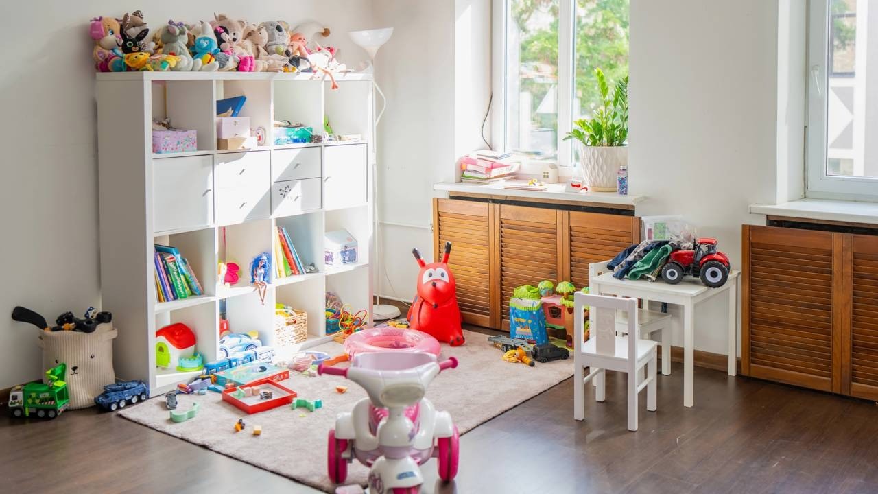 Quarto infantil desorganizado com brinquedos no chão, prateleira branca cheia de bichos de pelúcia e janela.