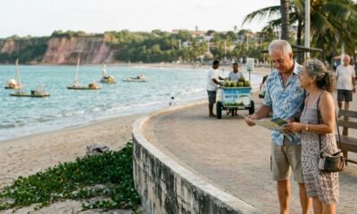 Casal idoso em calçadão, homem lendo mapa, mulher sorrindo. Vendedor de "LOCAL COCO WATER" na praia com barcos