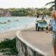Casal idoso em calçadão, homem lendo mapa, mulher sorrindo. Vendedor de "LOCAL COCO WATER" na praia com barcos