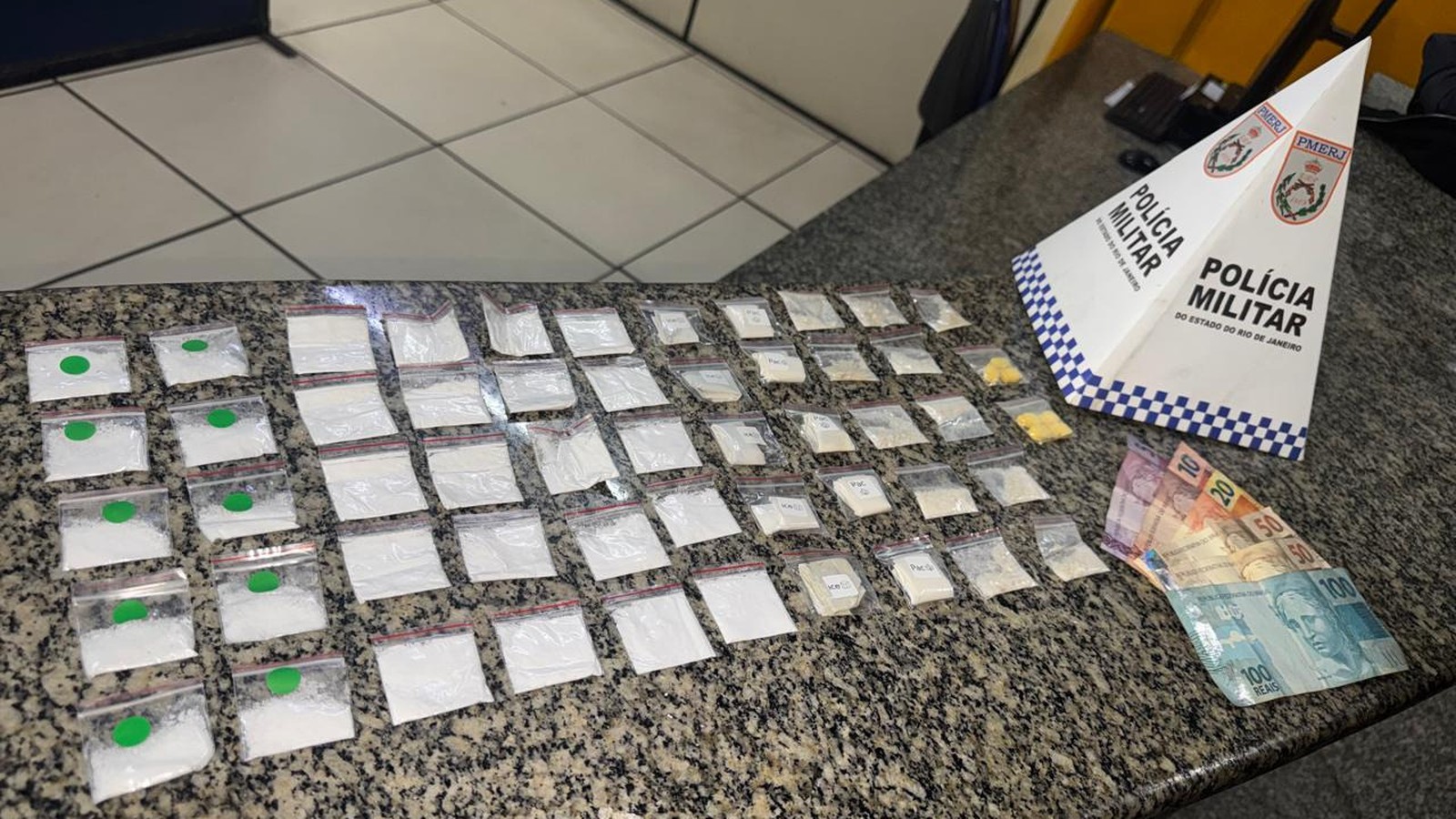 Drogas em sacolas, notas de dinheiro e placa da Polícia Militar sobre bancada