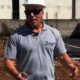 Homem de capacete branco, óculos escuros e camisa cinza com logo 'Henrique Projetos e Construção' em obra