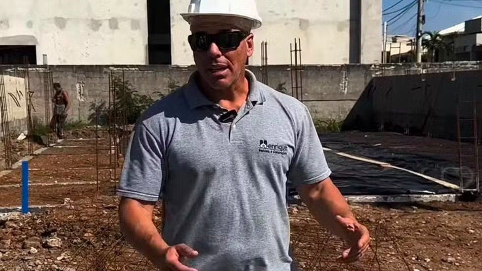 Homem de capacete branco, óculos escuros e camisa cinza com logo 'Henrique Projetos e Construção' em obra