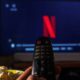 Mão segura controle remoto apontado para TV exibindo o logo Netflix, com pipoca